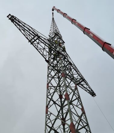 Grue GRIMM en action pour la construction d'une tour &eacute;lectrique haute tension avec des techniciens travaillant en hauteur.